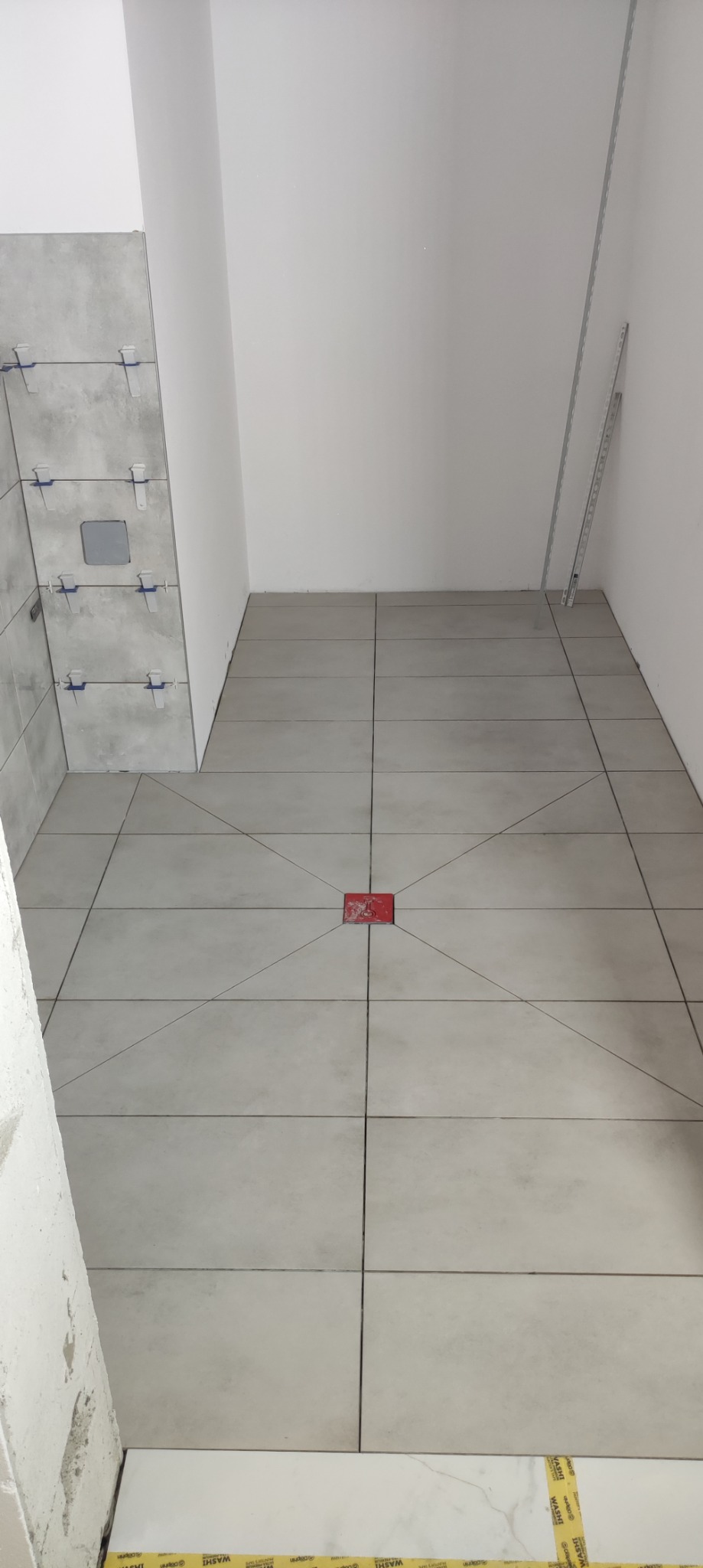 Podłoga w łazience w trakcie układania płytek, widoczne krzyżyki dystansowe na ścianie i wyznaczone linie cięcia na podłodze, czerwony odpływ liniowy w centrum.