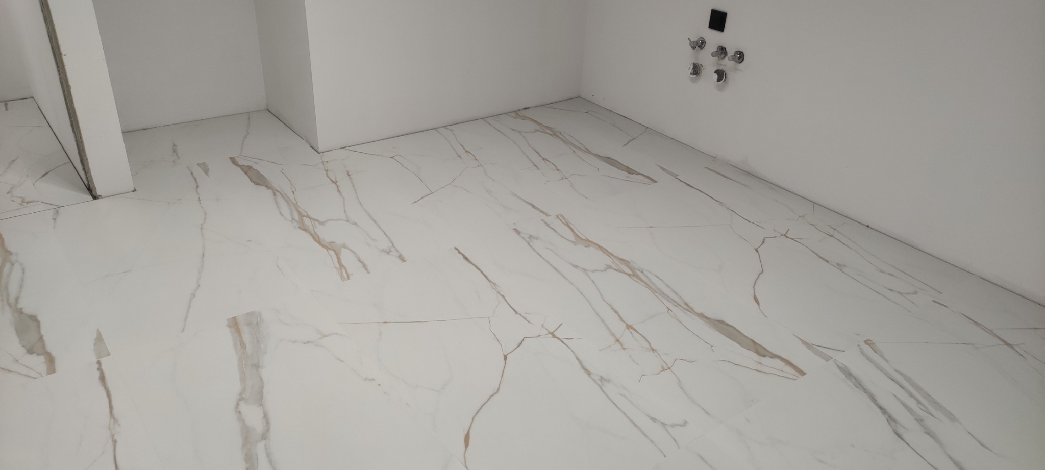 Nowo położona, błyszcząca podłoga z białych płytek imitujących marmur z wyraźnymi, złotymi żyłami, w minimalistycznej łazience z białymi ścianami i przygotowanymi przyłączami hydraulicznymi.