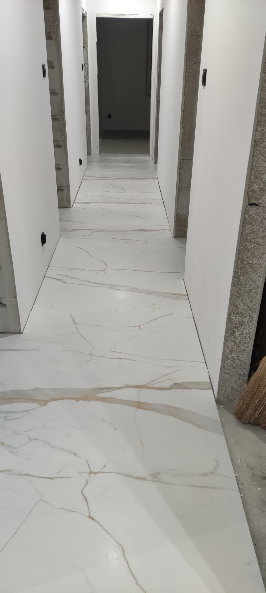 Korytarz w trakcie wykańczania, białe płytki podłogowe z marmurowym wzorem, surowe ściany z płyt kartonowo-gipsowych przed malowaniem.