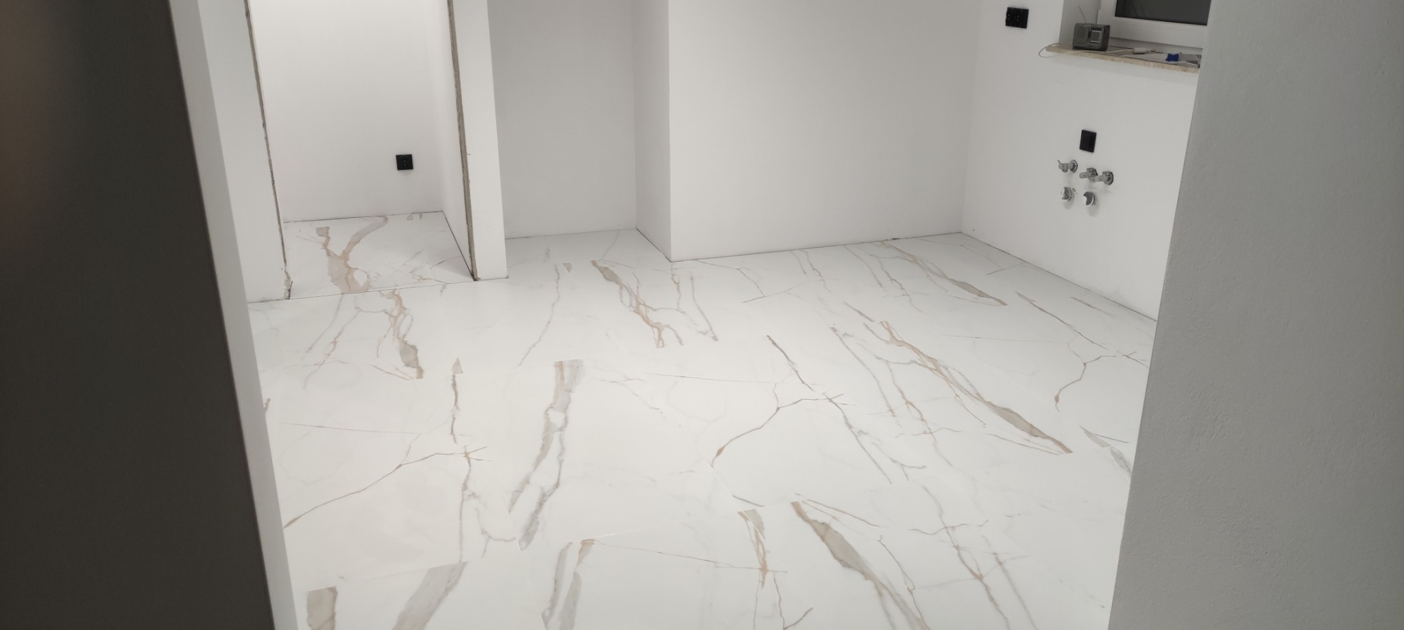 Wykończona podłoga w łazience z białych płytek imitujących marmur z delikatnymi szarymi żyłami, widoczne przygotowane przyłącza wodne i kanalizacyjne na ścianie, brak armatury.