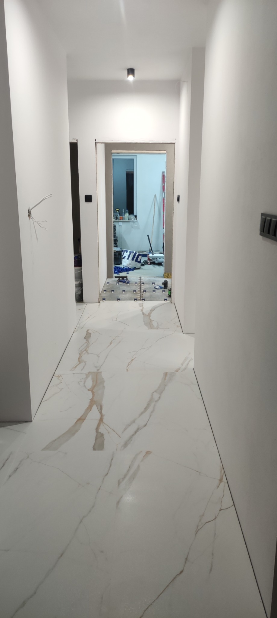 Korytarz w trakcie układania białych płytek imitujących marmur z brązowymi żyłami. Widoczne narzędzia glazurnicze i krzyżyki dystansowe.