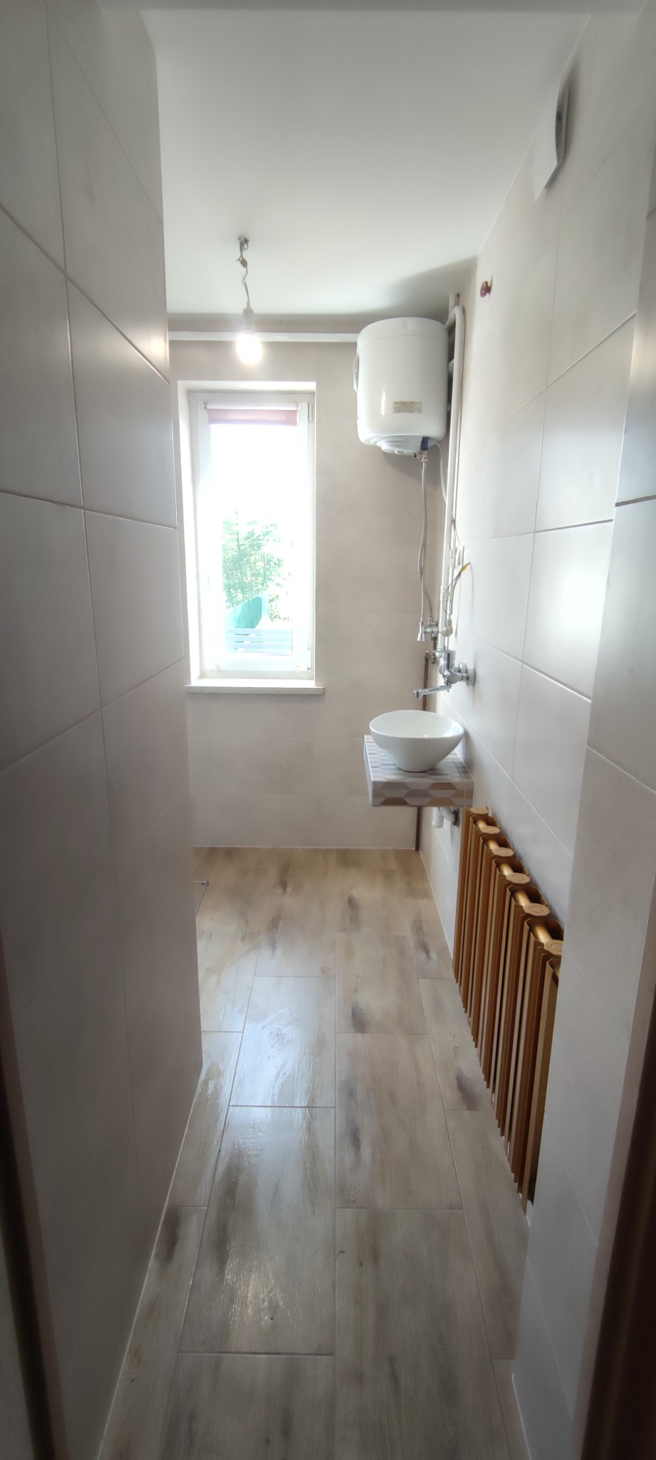 Wnętrze łazienki po remoncie: beżowe płytki na ścianach i podłodze imitujące drewno, biała umywalka na blacie z mozaiką, grzejnik drabinkowy z drewnianymi szczebelkami, bojler elektryczny...