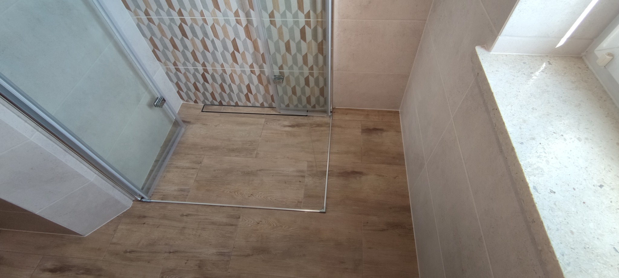 Nowoczesna kabina prysznicowa z geometrycznym wzorem płytek na ścianie, drewnopodobna podłoga, jasne płytki ścienne i parapet z lastryko.