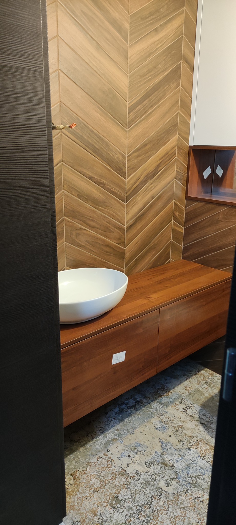 Łazienka z umywalką nablatową na drewnianej szafce, wzorzysta ściana z płytek ułożonych w jodełkę, widok zza uchylonych drzwi.