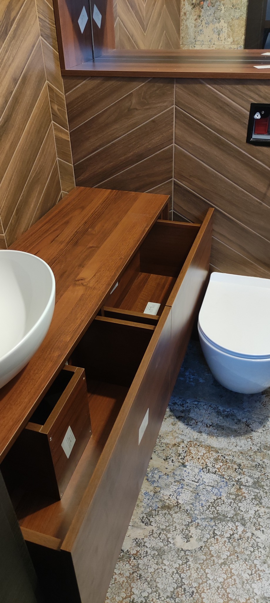 Nowoczesna łazienka z drewnianą szafką pod umywalką z otwartymi szufladami, białą umywalką nablatową i wiszącą toaletą, na podłodze wzorzyste płytki, ściany wyłożone płytkami imitującymi drewno...