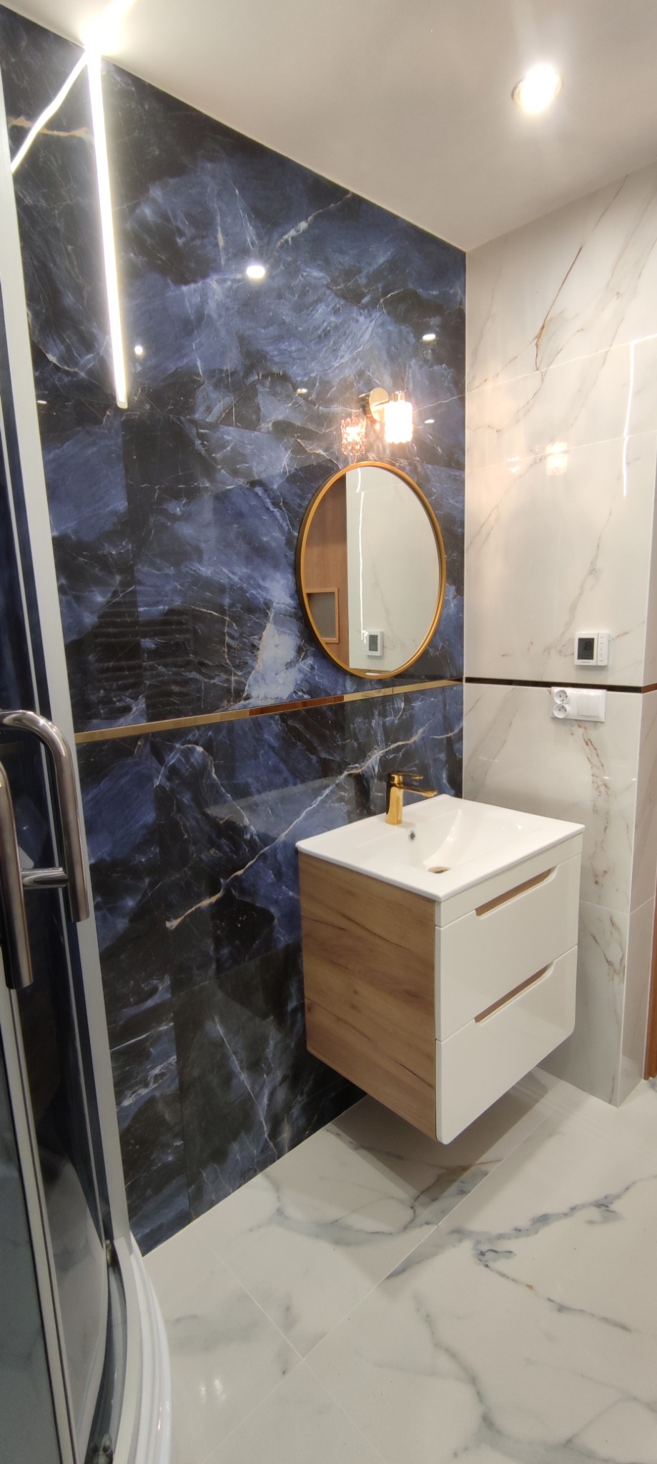 Nowoczesna łazienka z umywalką nablatową na szafce z drewnianymi frontami, okrągłym lustrem w złotej ramie i marmurowymi płytkami w odcieniach błękitu i bieli, oświetlenie LED.