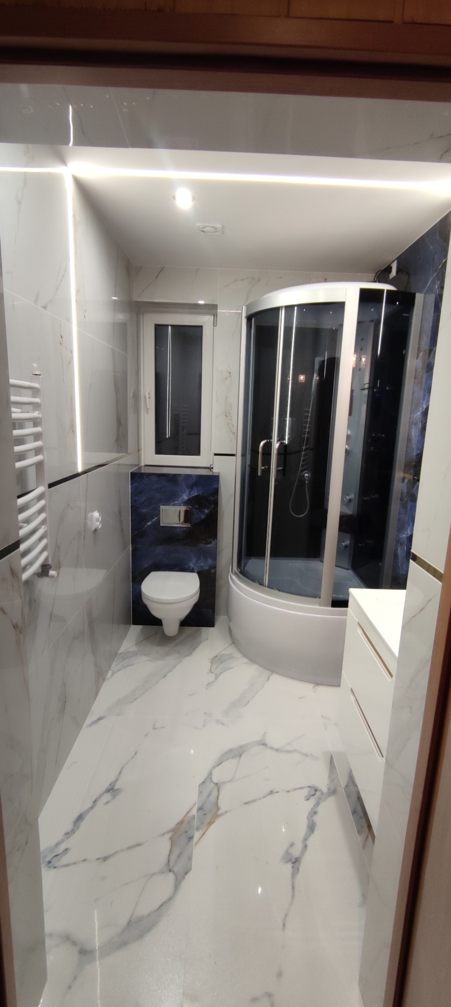 Nowoczesna łazienka z białą marmurową płytką, kabiną prysznicową z czarnymi akcentami, podwieszaną toaletą na tle niebieskiego marmuru i oświetleniem LED.