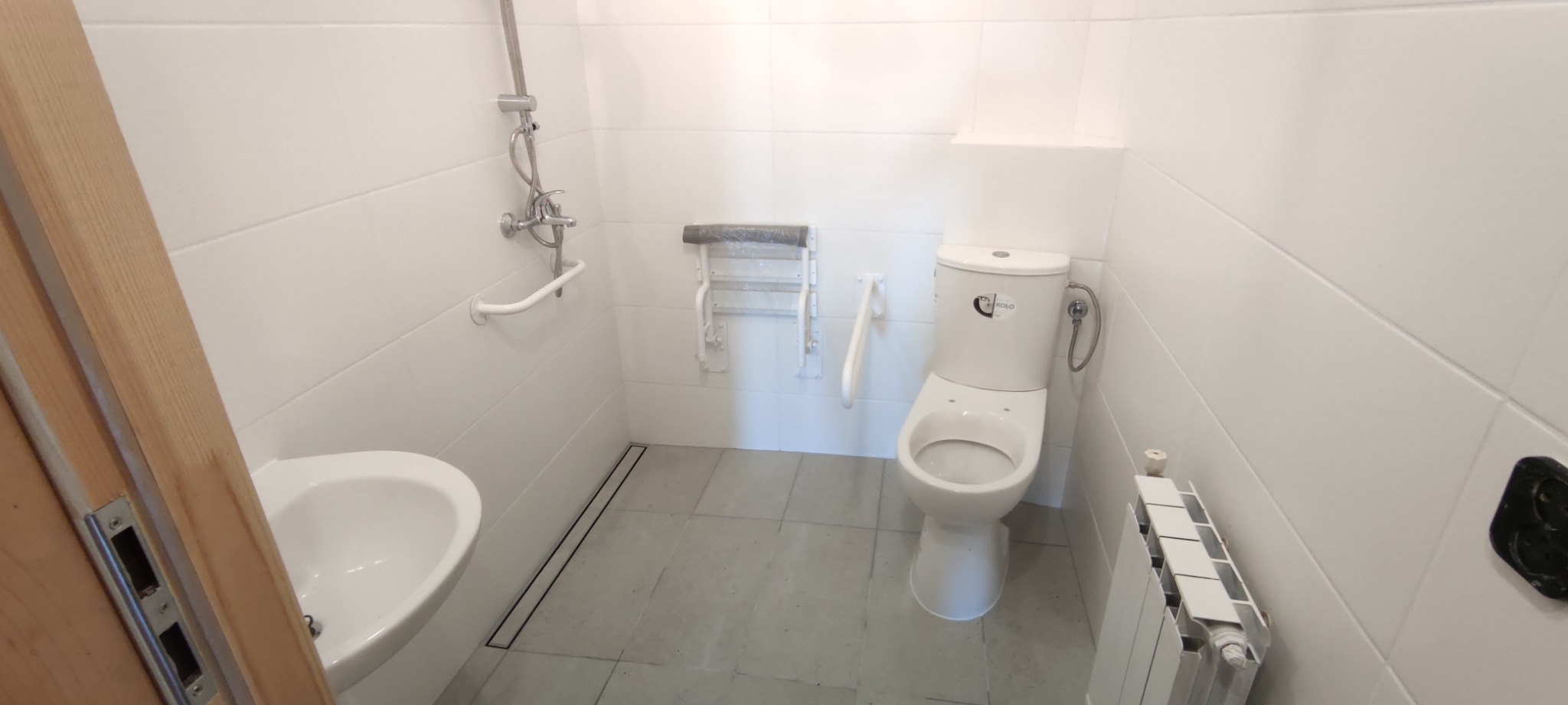 Jasna łazienka z białymi płytkami, umywalką, toaletą z bidetką, grzejnikiem, odpływem liniowym i składanym siedziskiem prysznicowym dla niepełnosprawnych.