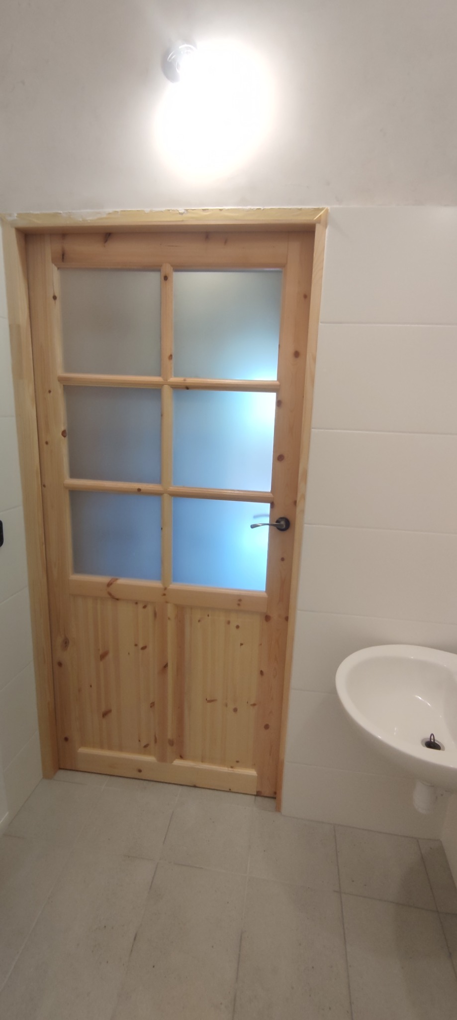 Jasna drewniana rama drzwi z matowym szkłem w łazience z białą umywalką i płytkami