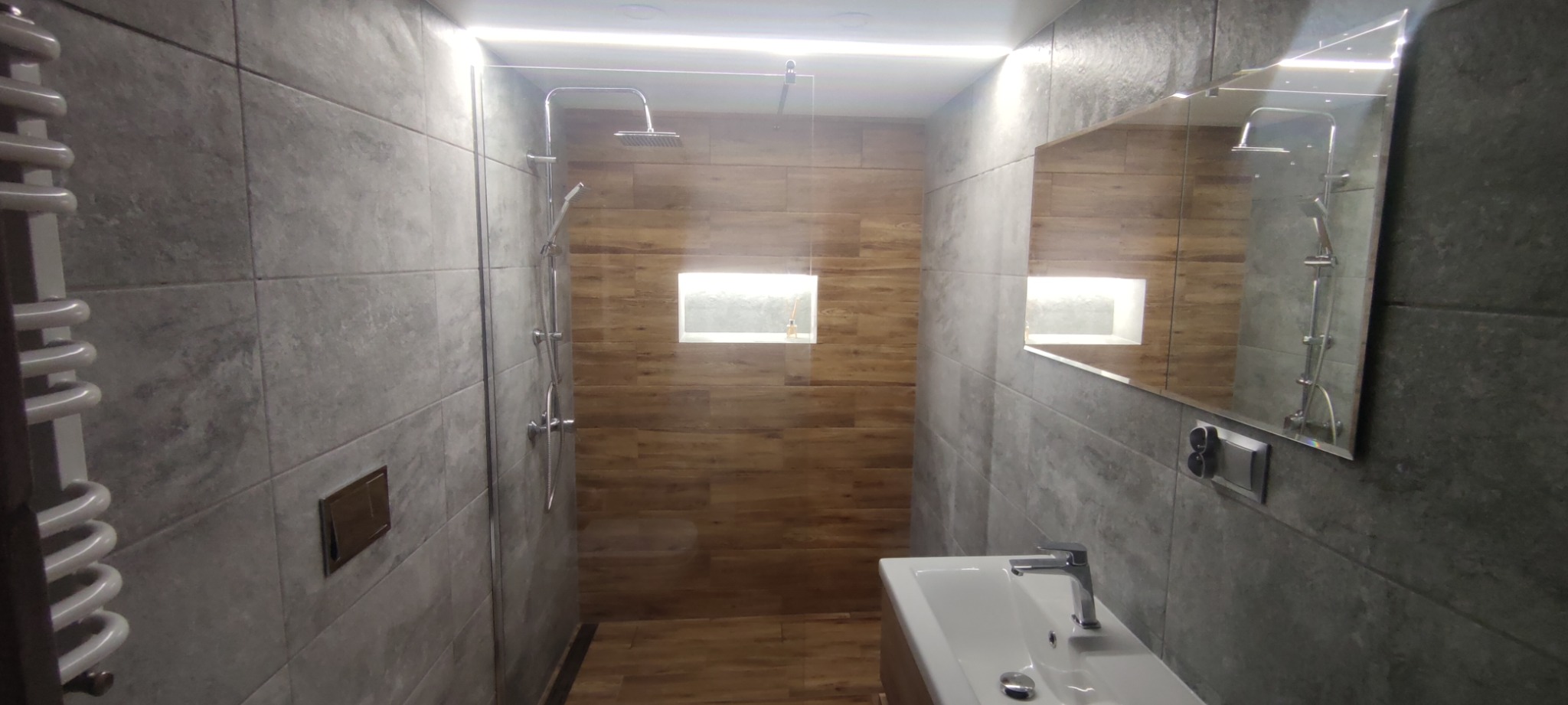 Nowoczesna łazienka z szarymi płytkami imitującymi beton, drewnopodobnymi płytkami pod prysznicem typu walk-in, podświetlaną wnęką i lustrem, widok na umywalkę z baterią.