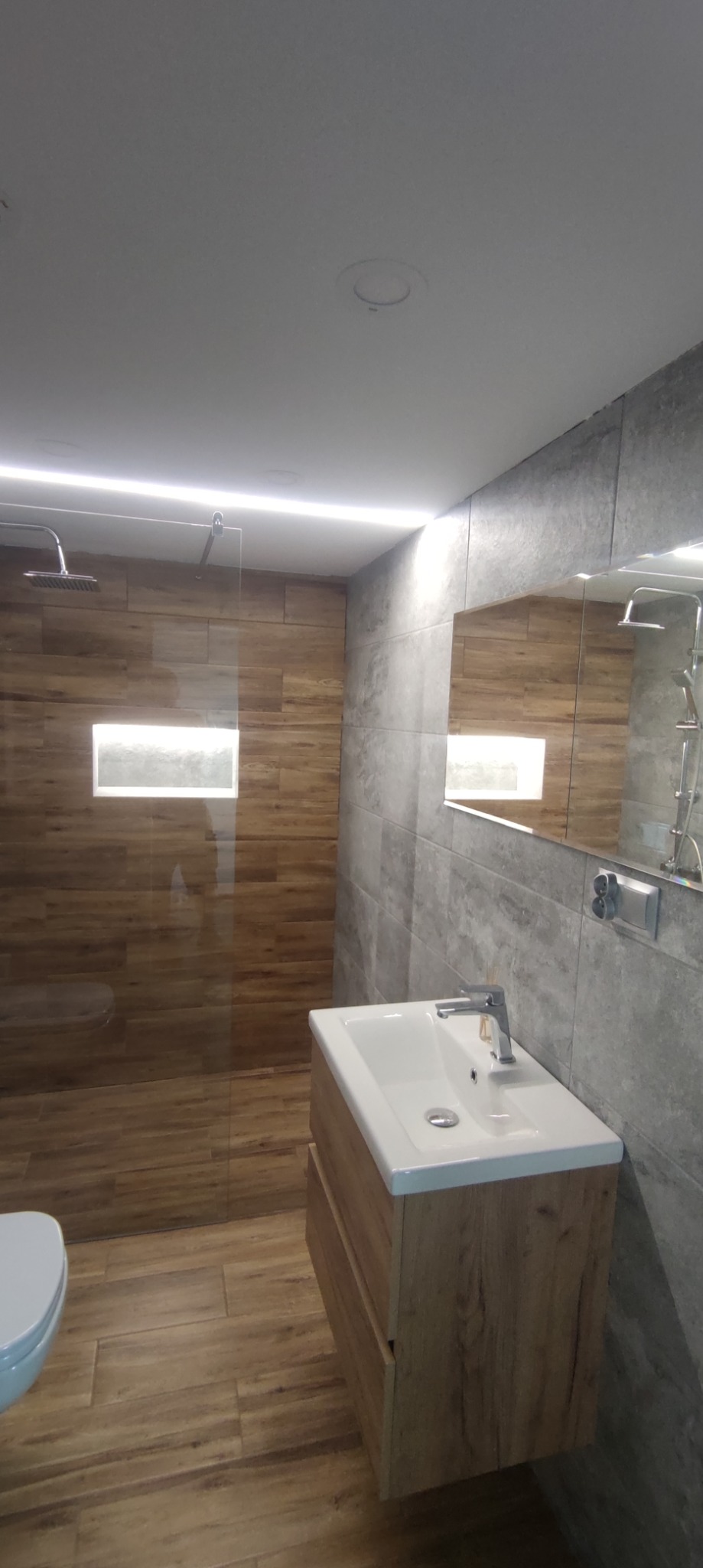 Nowoczesna łazienka z kabiną prysznicową typu walk-in, wykończona płytkami imitującymi drewno i szarością, oświetlenie LED podsufitowe, umywalka nablatowa z szafką w kolorze drewna.