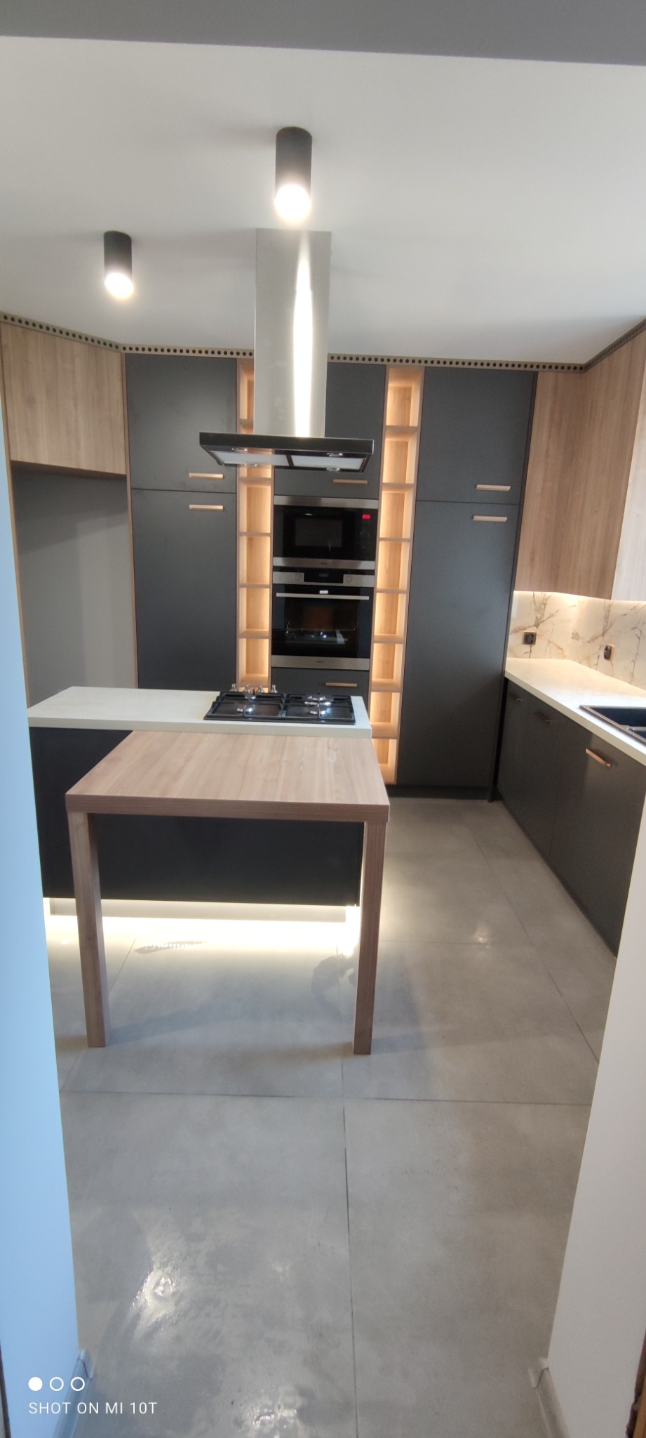 Nowoczesna kuchnia w odcieniach szarości i drewna z oświetleniem LED pod wyspą kuchenną i w regale obok piekarnika i mikrofalówki.