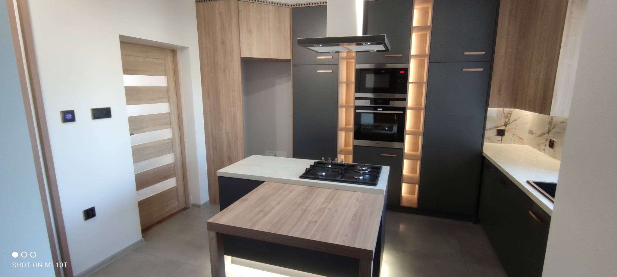 Nowoczesna kuchnia z wyspą, czarne szafki z drewnianymi akcentami, oświetlenie LED w półkach, sprzęt AGD w zabudowie, płyta gazowa na wyspie, drzwi z poziomymi paskami w tle.