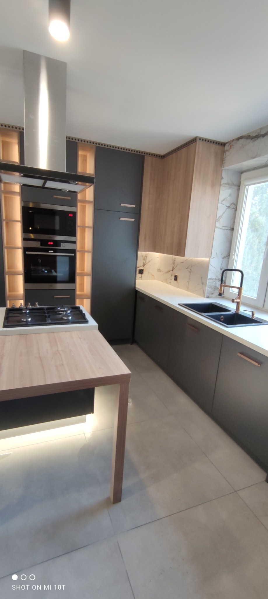 Nowoczesna kuchnia z szarymi szafkami, drewnianymi akcentami, marmurowym backsplash i wyspą z podświetleniem LED, widok z boku.