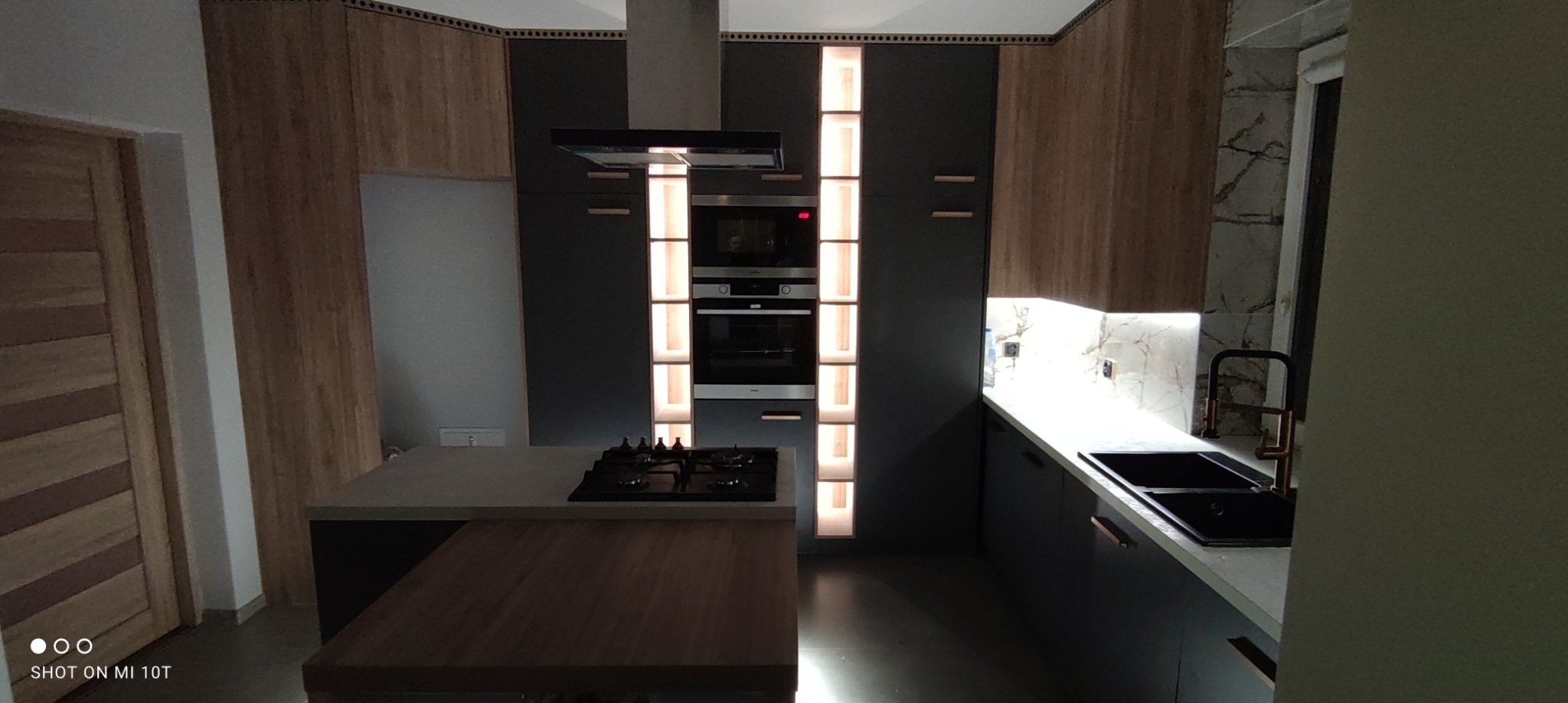 Nowoczesna kuchnia z wyspą, ciemnymi szafkami i drewnianymi akcentami, oświetlenie LED podkreśla pionowe linie zabudowy.