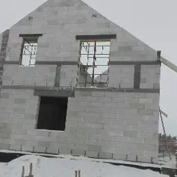 Surowy stan budynku z betonu komórkowego w zimowej scenerii, widoczne otwory okienne i fragment drewnianej konstrukcji dachu, na pierwszym planie śnieg.