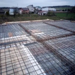 Zbrojenie płyty fundamentowej z widocznymi deskami szalunkowymi i izolacją, w tle zabudowa jednorodzinna pod pochmurnym niebem.