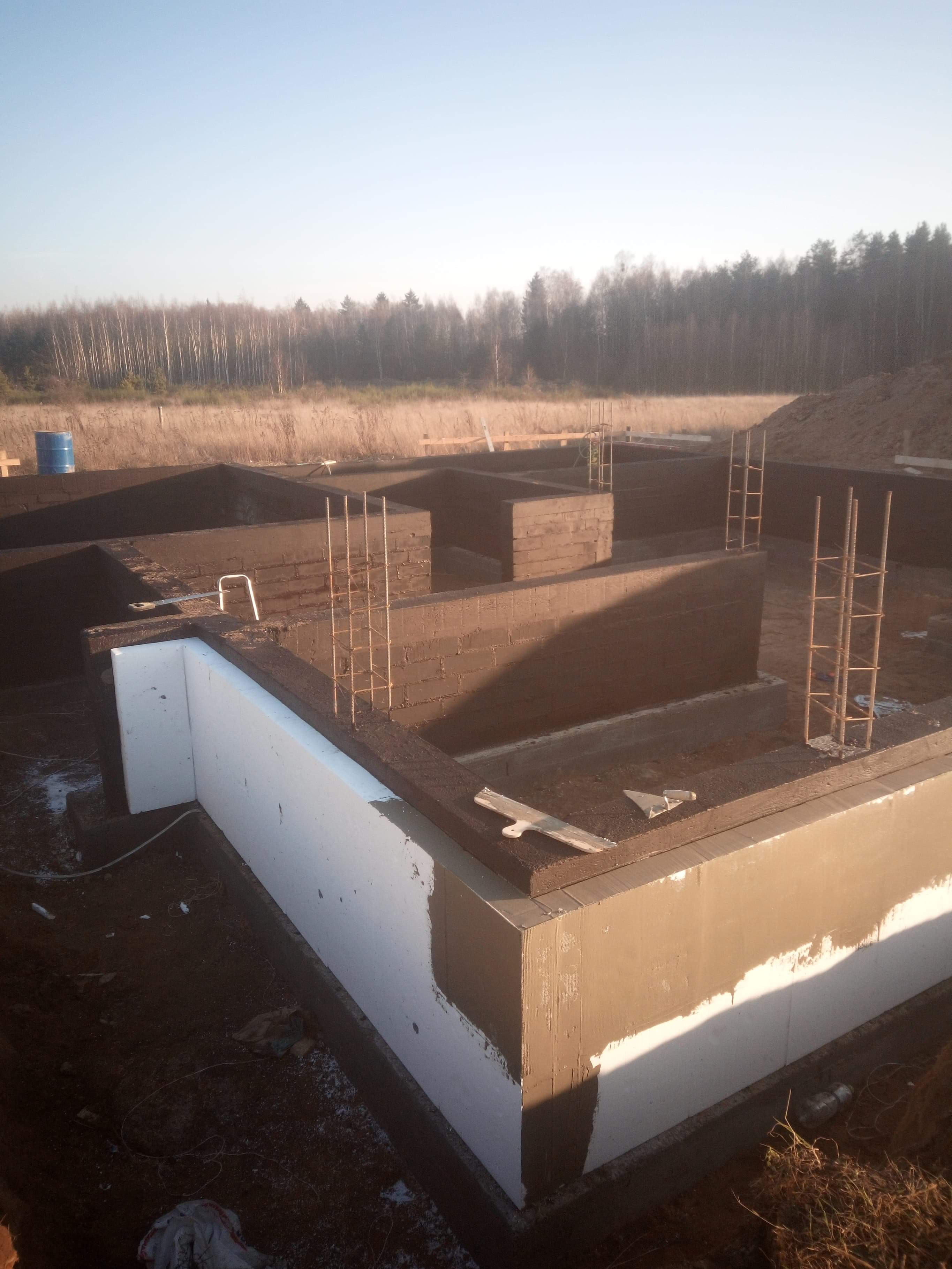 Zalane fundamenty z izolacją termiczną, widoczne zbrojenia pionowe i narzędzie murarskie, w tle las.