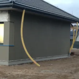 Fragment elewacji domu jednorodzinnego z szarą strukturą tynku, widoczny fragment dachu z szarych dach&oacute;wek oraz ż&oacute;łte rury wentylacyjne przy ścianie.