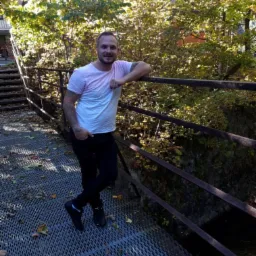 Mężczyzna opiera się o rdzawą barierkę mostu z metalowej kraty, w tle jesienne drzewa i budynek. Widoczny tatuaż na ramieniu i uśmiechnięta twarz.