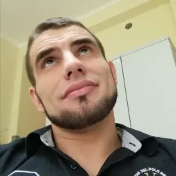 Mężczyzna z brodą spogląda w górę na sufit z kratką wentylacyjną i białą szafką w tle.