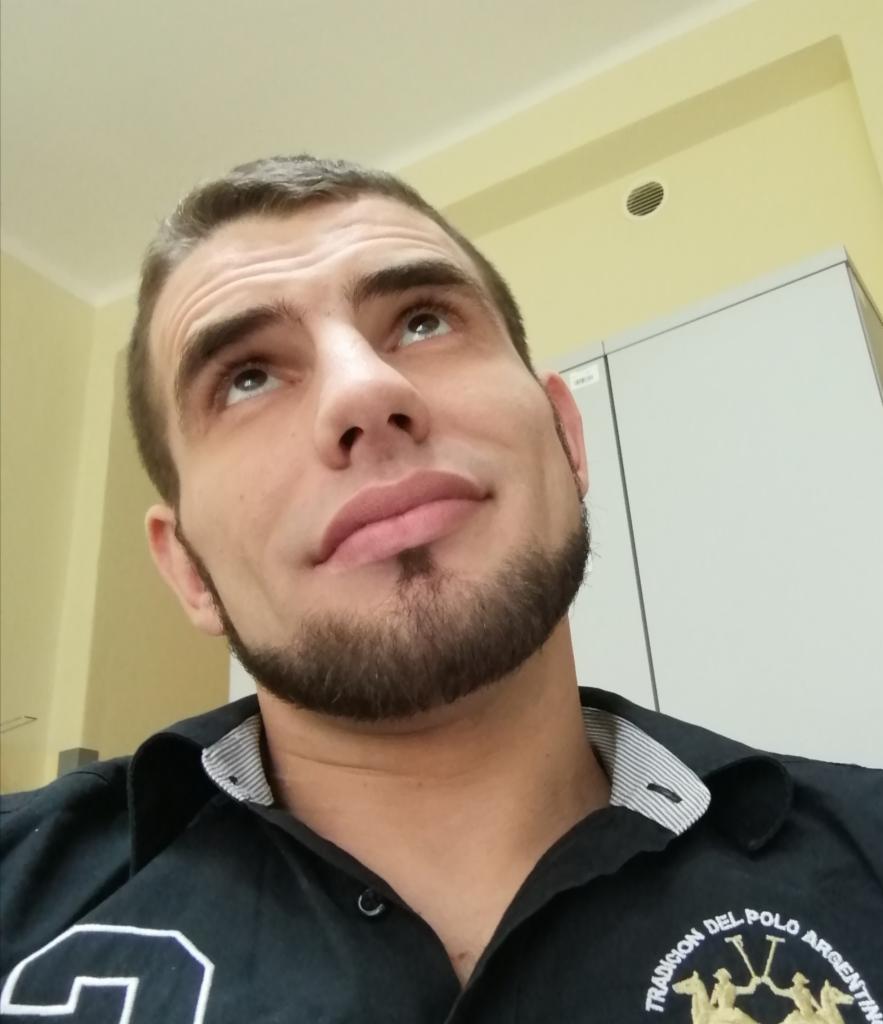 Mężczyzna z brodą spogląda w górę na sufit z kratką wentylacyjną i białą szafką w tle.