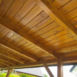 Drewniana konstrukcja z belek i desek, prawdopodobnie zadaszenie tarasu lub altany, widok od dołu, z widocznymi elementami krajobrazu w tle.
