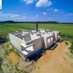 Surowy stan domu z keramzytu w trakcie budowy, widok z lotu ptaka na fundamenty i ściany, z zielonym polem w tle.