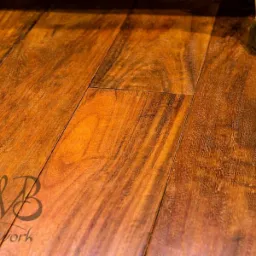 Połyskująca, świeżo polakierowana podłoga z ciemnego drewna o wyraźnym usłojeniu, z fragmentem złotej nóżki mebla w prawym górnym rogu i logo firmy na pierwszym planie.