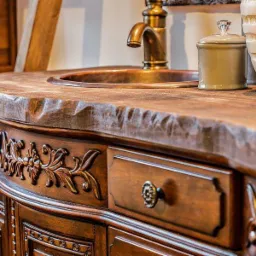 Rustykalna umywalka z miedzianą baterią i drewnianym blatem o nieregularnej krawędzi, zdobiona szafka z rzeźbionymi motywami roślinnymi i ceramiczne akcesoria.
