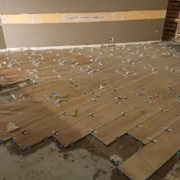 Układanie podłogi z paneli imitujących drewno, z użyciem niebieskich klipsów poziomujących i czarnego wiadra na klej w rogu pomieszczenia.