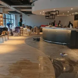 Wnętrze lokalu gastronomicznego w trakcie prac wykończeniowych: układanie podłogi, montaż baru i regałów, widoczne narzędzia, materiały budowlane i ekipa remontowa.