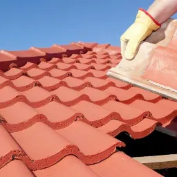 Czerwona dachówka ceramiczna układana na dachu, widoczne ręce w żółtych rękawicach i fragment buta roboczego, błękitne niebo w tle.