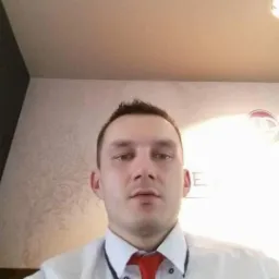 Portret mężczyzny w białej koszuli i czerwonym krawacie na tle ściany z delikatnym wzorem i sufitu z lampą.