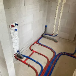 Surowy stan łazienki z widocznymi rurami instalacji wodnej (czerwone i niebieskie) przymocowanymi do podłoża, pionowe rury wychodzące ze ściany zabezpieczone pianką montażową, ściany z białych...