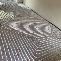 Białe rurki grzewcze ułożone spiralnie na srebrnej folii z czerwoną kratką, tworzące system ogrzewania podłogowego w trakcie instalacji.