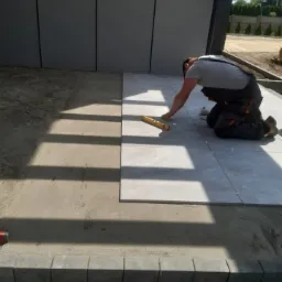 Układanie jasnych płytek na zewnątrz budynku, widoczny pracownik z poziomicą sprawdzający równość, przygotowane podłoże z piasku.