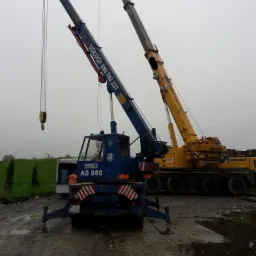 Dwa dźwigi kołowe na placu budowy: niebieski dźwig z numerem AD 080 i żółty dźwig Liebherr w tle, pochmurny dzień.