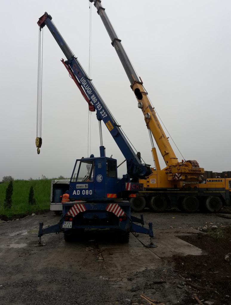 Dwa dźwigi kołowe na placu budowy: niebieski dźwig z numerem AD 080 i żółty dźwig Liebherr w tle, pochmurny dzień.