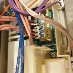 Wnętrze pralki z widocznymi kablami różnej grubości i kolorów, złącza elektryczne oraz płyta drukowana, pokryte warstwą kurzu.