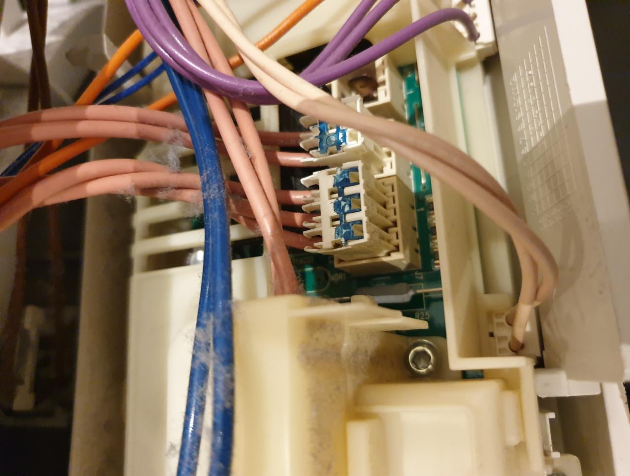 Wnętrze pralki z widocznymi kablami różnej grubości i kolorów, złącza elektryczne oraz płyta drukowana, pokryte warstwą kurzu.