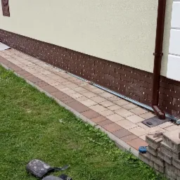 Montaż brązowej okładziny z płytek klinkierowych na cokole budynku, widoczne krzyżyki dystansowe, obok ułożona kostka brukowa w dwóch odcieniach, rynna spustowa, trawnik.