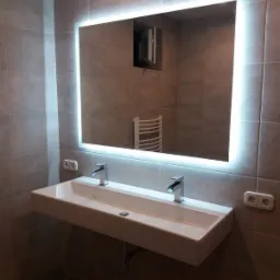 Nowoczesna łazienka z podwójną umywalką nablatową, oświetlonym lustrem i szarą glazurą na ścianach. W lustrze odbija się biały grzejnik drabinkowy.