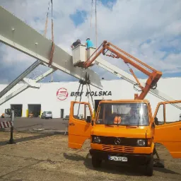 Pomarańczowy podnośnik koszowy Mercedes-Benz z otwartymi drzwiami kabiny unosi stalową konstrukcję na tle budynku BMF Polska i pochmurnego nieba.