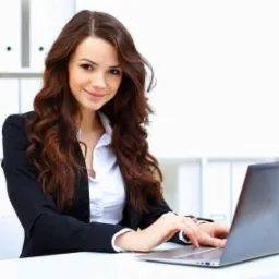 Uśmiechnięta kobieta w czarnej marynarce i białej koszuli, siedząca przy biurku i pisząca na laptopie; w tle białe półki z segregatorami.