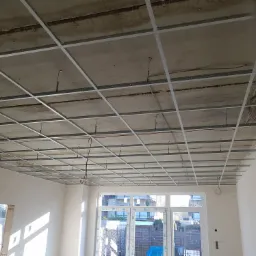 Montaż metalowej konstrukcji podwieszanego sufitu w surowym pomieszczeniu z widocznymi przewodami elektrycznymi i oknem z widokiem na zabudowę jednorodzinną.