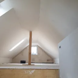 Poddasze w trakcie prac wykończeniowych z widoczną konstrukcją wspornikową, oknem dachowym i ścianami przygotowanymi pod malowanie.
