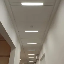 Długi korytarz biurowy z podwieszanym sufitem kasetonowym i liniowym oświetleniem LED, widoczne kratki wentylacyjne w suficie oraz klimatyzatory na ścianie.