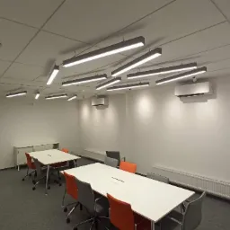 Nowoczesna sala konferencyjna z podwieszanymi, liniowymi lampami LED, białymi stołami i krzesłami w kolorach szarym i pomarańczowym, klimatyzatory na ścianach.