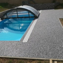 Royal Floor - Wykończony teren wokół basenu z szarą nawierzchnią z drobnych kamyczków, częściowo zadaszony basen z błękitną wodą, w tle trawnik i kamienny murek.